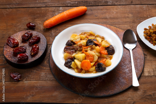 A dish of Jewish cuisine - tsimes with carrots and dates in a white plate on a round stand next to a spoon and carrots with dried fruits. photo