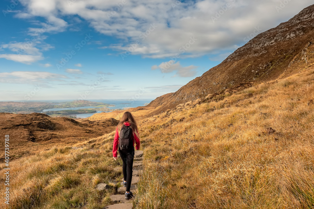 Teenager girl walking on a path by a mountain. Beautiful scenery in the background. Connemara National park, county Galway, Ireland. Warm sunny day, Cloudy sky. Outdoor activity and tourism concept