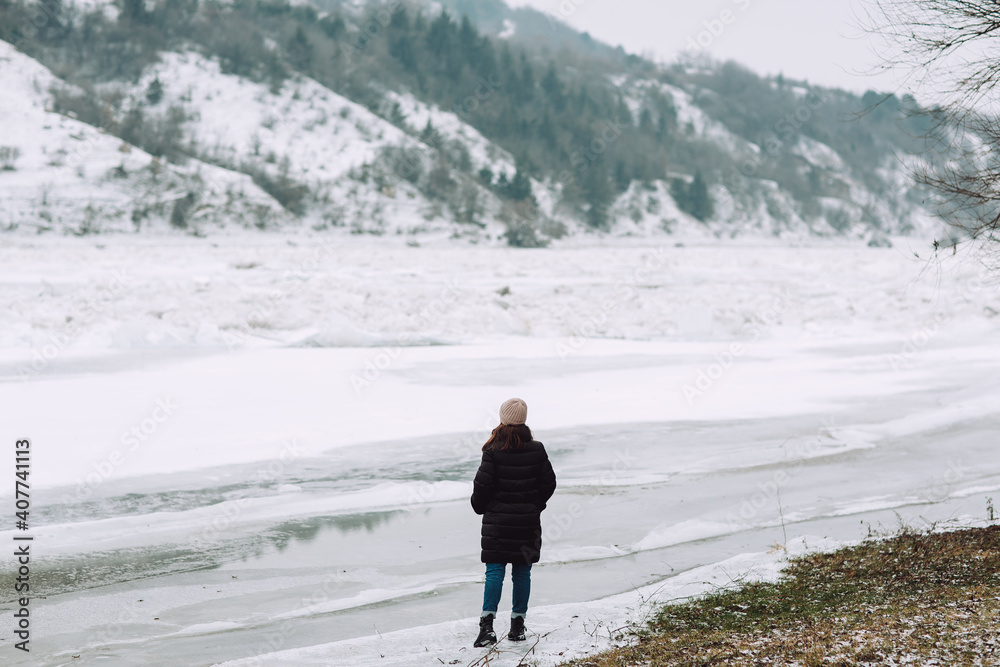 Winter landscape. One girl stands in front of a winter snowy river and looking to the beautiful landscape.