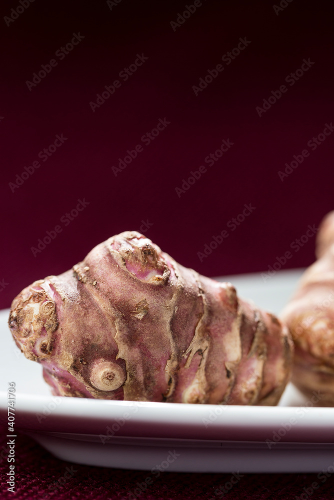 Detail of single Jerusalem artichokes (Helianthus tuberosus), also known as sunroot, sunchoke, or earth apple, lay on a white plate against a scarlet background. Vertical frontal view with text space Detail of single Jerusalem artichokes (Helianthus tuberosus), also known as sunroot, sunchoke, or earth apple, lay on a white plate against a scarlet background. Vertical frontal view with text space