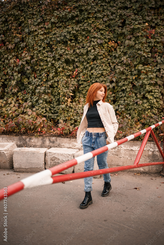 Fototapeta premium A pretty redhead young woman is walking down the street, she is dressed in jeans and a beige shirt. Beautiful girl dressed in casual style with a smile on her face