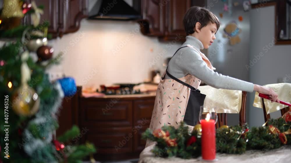 Side view of little Caucasian brunette boy lightning candles on Christmas table indoors. Happy child in apron preparing New Year dinner for family at home in kitchen. Holiday celebration and joy. Side view of little Caucasian brunette boy lightning candles on Christmas table indoors. Happy child in apron preparing New Year dinner for family at home in kitchen. Holiday celebration and joy.