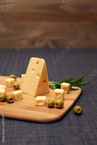 A piece of cheese with holes and olives on a wooden board. Maasdam cheese.