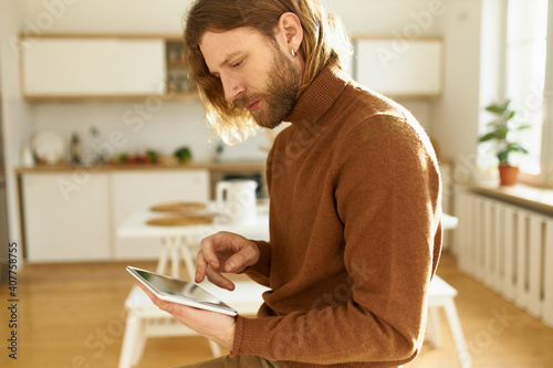 Profile image of concentrated young redheaded man with earring and pierced nose using wireless high speed internet connection on digital tablet, playing new video game via online application