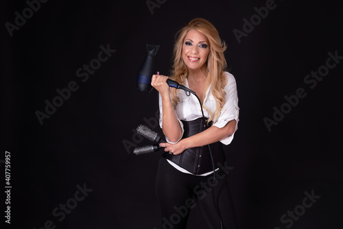 Blonde hairdresser holding a hairdryer and hair brushes while smiling on a black background.