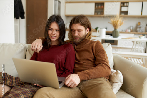 Cute young couple bonding at home. Unshaven male hugging his pretty girlfriend, sitting in living room with portable computer on lap, enjoying nice time together, browsing internet on weekend