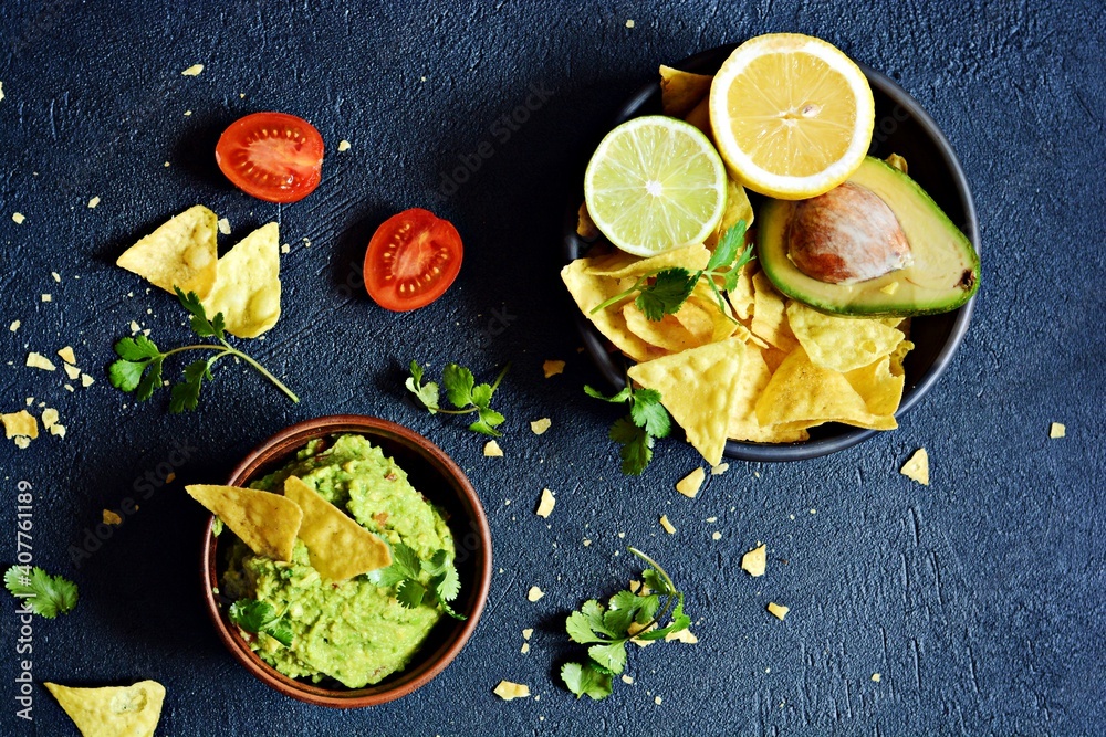 Bowl of guacamole dip with corn  nachos (chips) and ingredients on dark background, selective focus. Mexican national dish.