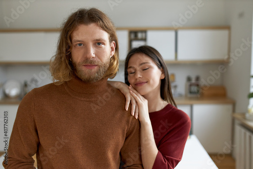 Portrait of fashionable young unshaven man with loose red hair posing indoors in stylish kitchen, cute girlfriend placing hands on his shoulder, keeping eyes closed, enjoying sweet moment together