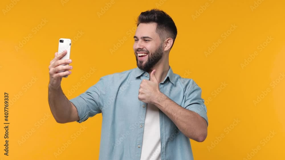 Cheerful bearded young man 20s years old in blue shirt isolated on yellow background studio. People lifestyle concept. Doing selfie shot on mobile phone showing biceps muscles victory sign thumb up Cheerful bearded young man 20s years old in blue shirt isolated on yellow background studio. People lifestyle concept. Doing selfie shot on mobile phone showing biceps muscles victory sign thumb up