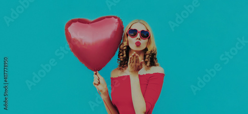 Portrait of beautiful young woman with red heart shaped balloon blowing her lips sending sweet air kiss on a blue background