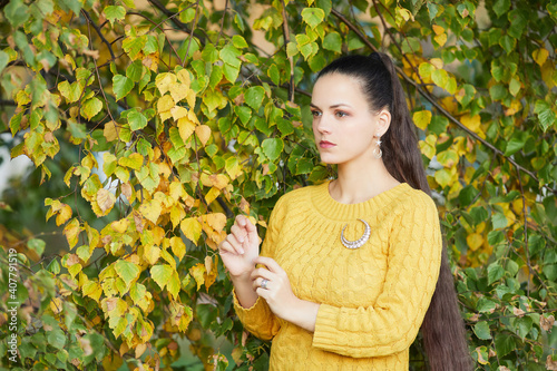 Portrait of a beautiful authentic young Russian woman in stylish cozy orange clothing. Girl posing against a background of yellow autumn leaves. Girl's facial skin is white, clean, and free of pimples