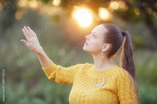 Young woman dressed in orange enjoys a sunset stroll and looks out into the distance. Behind the girl is a blurry forest and the rays of the setting sun