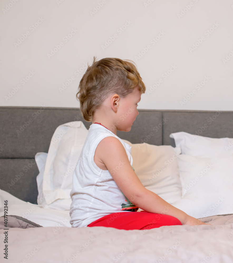 Offended little boy sitting on the bed in the bedroom. Upset boy in pajamas sitting in the bedroom on the bed. Offended little boy sitting on the bed in the bedroom. Upset boy in pajamas sitting in the bedroom on the bed.