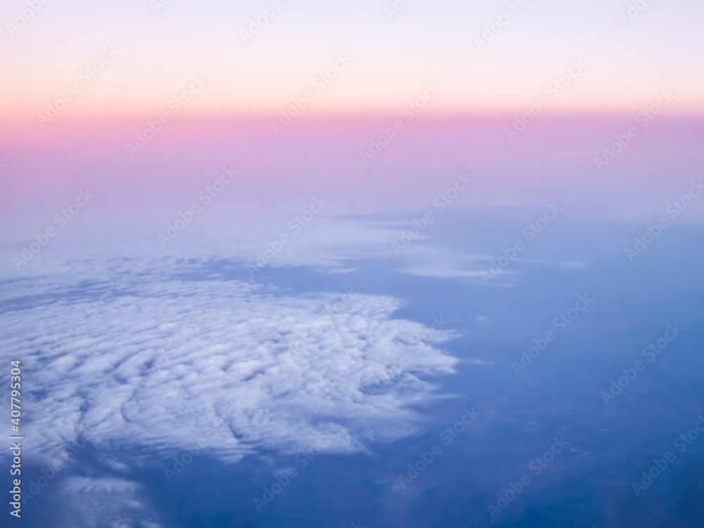 Clouds are viewed from above in flight at high altitude as the sunrise colors the sky at the horizon.