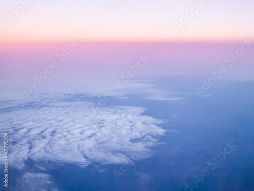 Clouds are viewed from above in flight at high altitude as the sunrise colors the sky at the horizon.