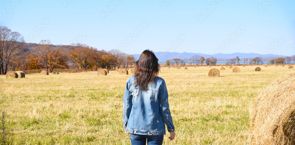 Young brunette woman in denim jacket standing near haystack in field with bales of hay. she looking at mountains and the autumn forest. Young brunette woman in denim jacket standing near haystack in field with bales of hay. she looking at mountains and the autumn forest.