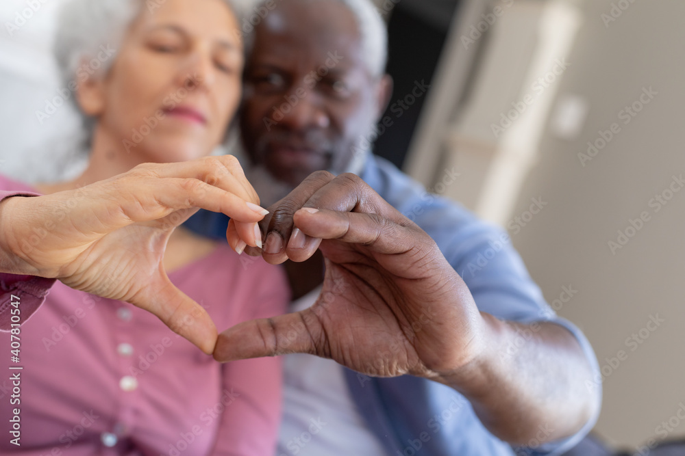 Senior mixed race couple in living room embracing making a heart shape with their hands Senior mixed race couple in living room embracing making a heart shape with their hands