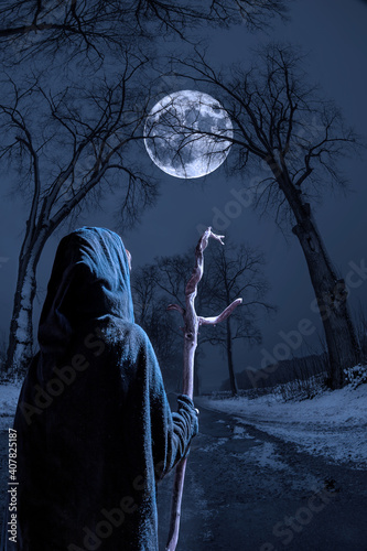 A witch with a stick walks down a narrow street on a winter night. A big full moon shines through tall trees.