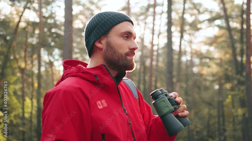 Close up of young handsome caucasian man in hat and red jacket standing in park and enjoying view looking through binoculars. Close up of young handsome caucasian man in hat and red jacket standing in park and enjoying view looking through binoculars.