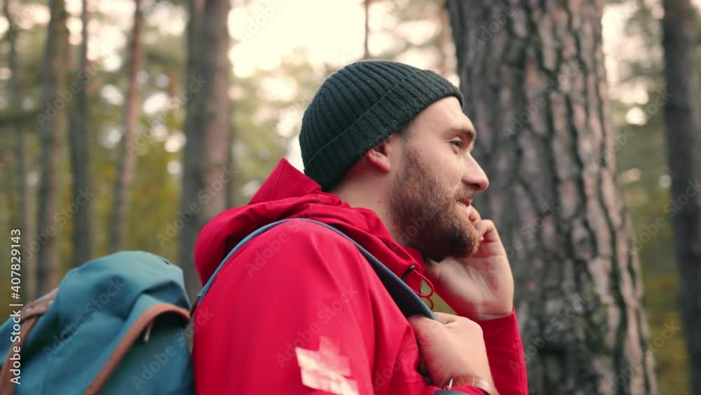 Close up of young caucasian bearded man talking on cell phone while standing in woods and looking around. Close up of young caucasian bearded man talking on cell phone while standing in woods and looking around.