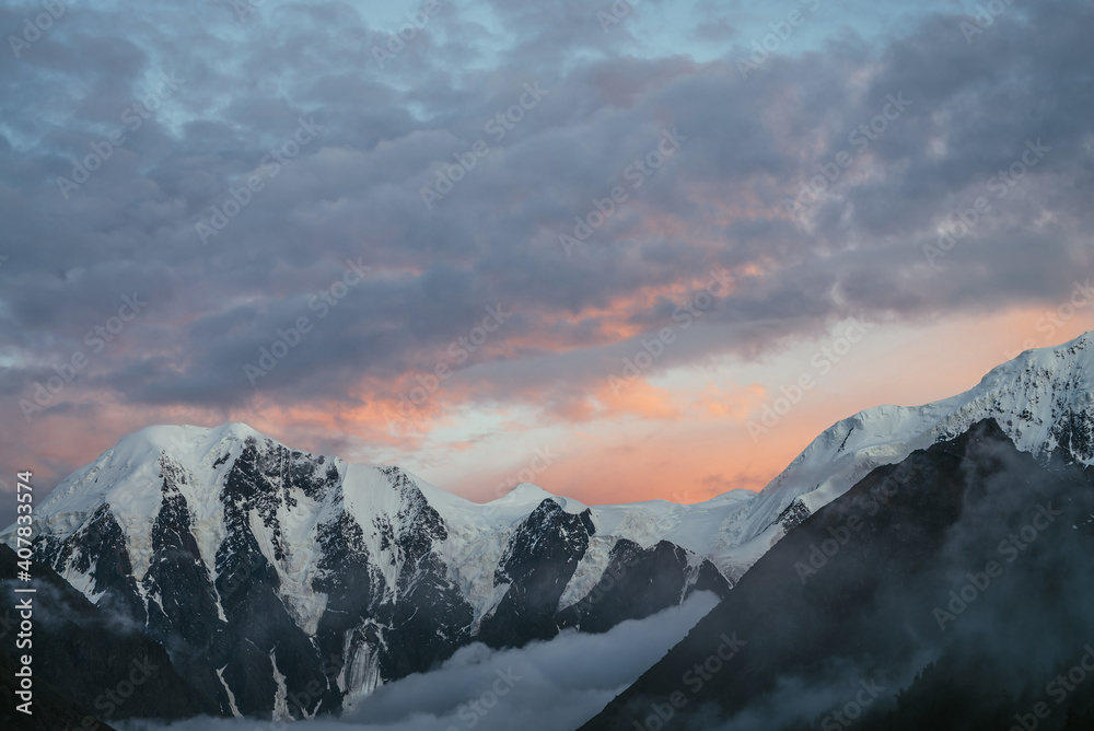 Atmospheric mountain landscape with great snowy mountains and low clouds in valley under violet orange dawn cloudy sky. Awesome alpine scenery with dense fog in mountain valley at sunset or at sunrise
