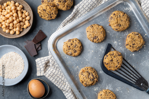 Horizontal composition of a baking tray with freshly baked chickpea cookies and dark chocolate chips, some ingredients and a turner