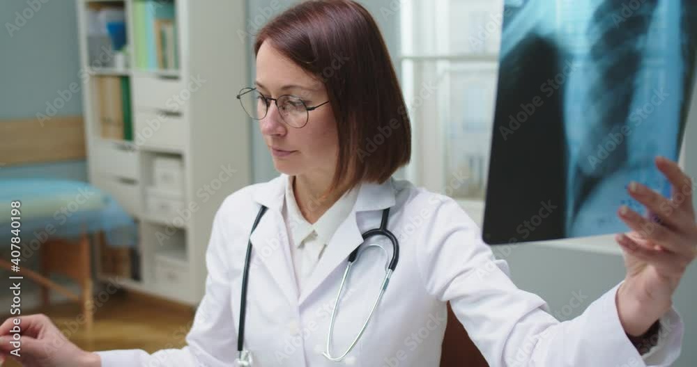 Portrait of beautiful Caucasian woman in glasses working in hospital. Adult female doctor in white medical gown looking at x-ray scan and making reports on computer. Medicine, health concept. Portrait of beautiful Caucasian woman in glasses working in hospital. Adult female doctor in white medical gown looking at x-ray scan and making reports on computer. Medicine, health concept.