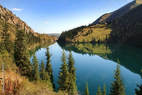 Mountain lake Kolsay surrounded by trees. Sunny summer day on an alpine lake, the lake between the mountains goes far away, trees, grass and bushes grow on the slopes of the mountains, reflections