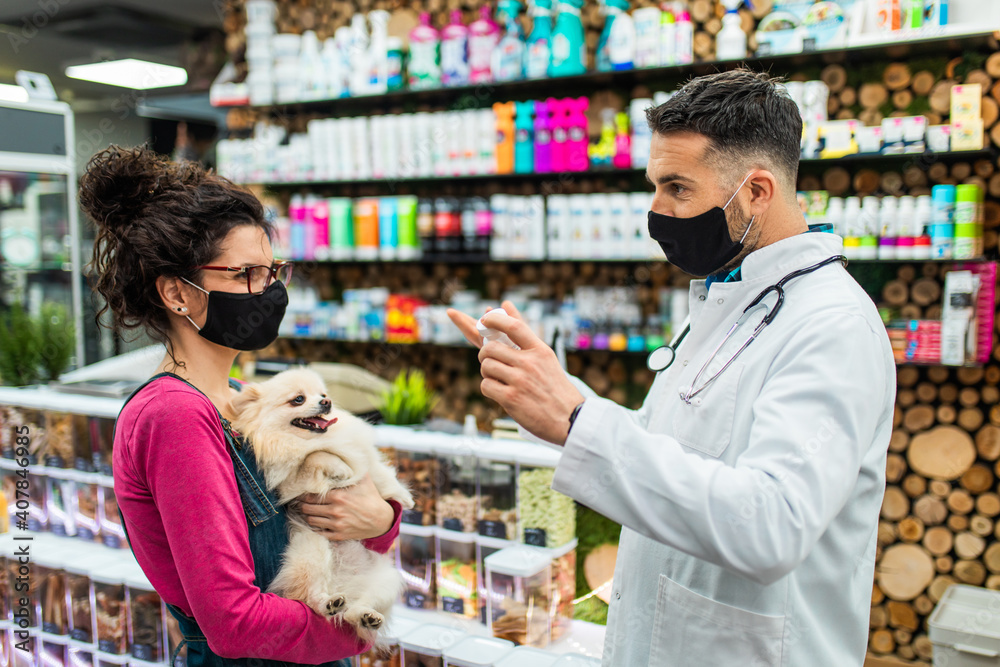 Female customer with protective face mask talking with veterinarian in pet shop and holding cute Pomeranian dog.