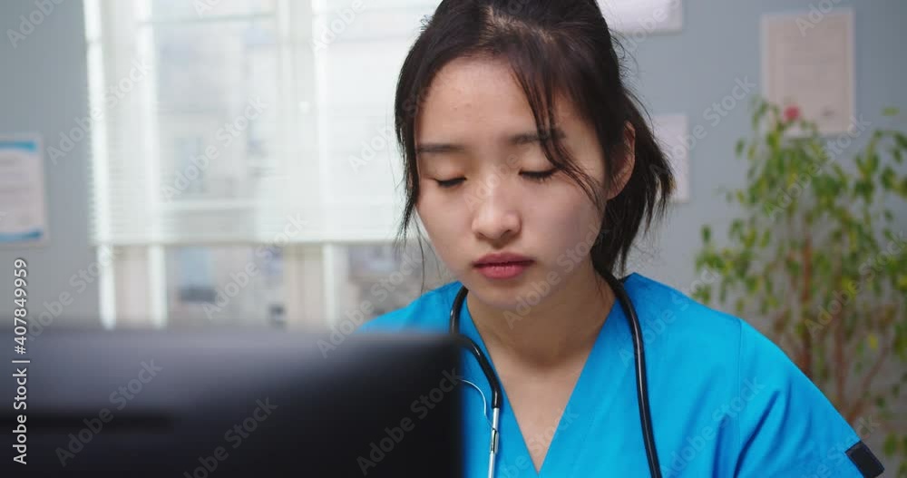 Portrait of attractive female nurse in blue medical uniform working on computer. Young Asian woman with stethoscope making e-reports, typing and checking documents in hospital. Medicine concept. Portrait of attractive female nurse in blue medical uniform working on computer. Young Asian woman with stethoscope making e-reports, typing and checking documents in hospital. Medicine concept.