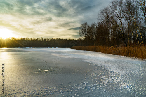 Frozen Pogoria II Lake with ice on it and with a sun behind clouds over a line of trees in a horizon.