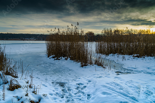 Frozen fishing place in winter at the Pogoria II Lake with heavy clouds and sun over horizon.