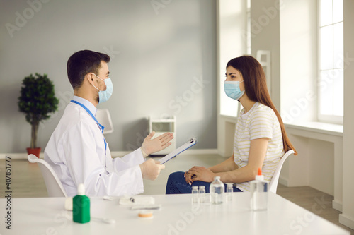 Doctor talking to patient during immunization and vaccination campaign at the clinic or hospital. Medical specialist in face mask interviewing young woman before injecting her with antiviral vaccine