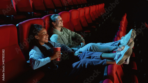 Fotografia Joyful young couple of friends laughing, putting their feet on seats while watch