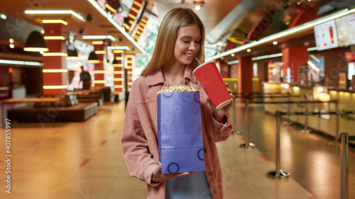 Portrait of happy young woman holding big popcorn bag and soda while posing in front of cinema bar in a movie theater
