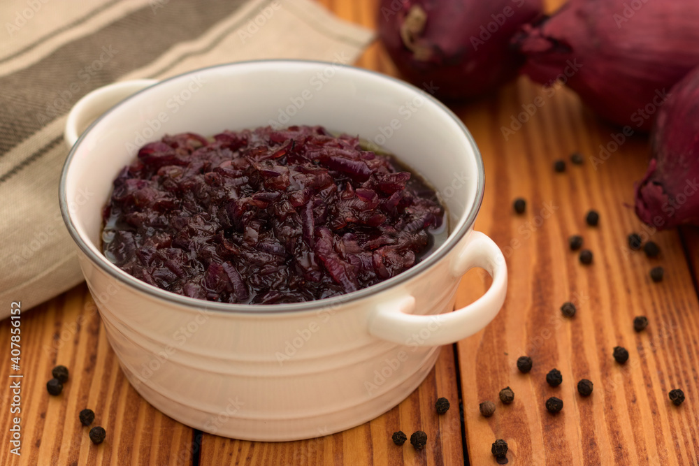 Red onion relish homemade with balsamic vinegar and brown sugar in a white ceramic dish standing on a wooden background. Red onion relish homemade with balsamic vinegar and brown sugar in a white ceramic dish standing on a wooden background.