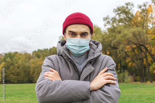 Man in a medical mask stands in the open air, a serious guy with his hands folded on his chest, portrait