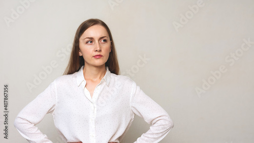 Suspicious young woman in a white shirt, hands on the waist, white background, copy space, 16:9 © Анастасия Семашко