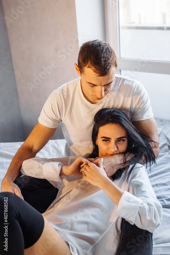 Young heterosexual couple of young lovers indulge in bed at home. Girl lies on legs of boyfriend and hugging his face with hand. Dressed in white shirts and dark pants. Pure enjoyment of each other © Daria