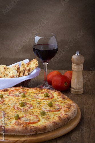 Photograph of a delicious Mozzarella Cheese Pizza with slices of red tomatoes and green olives. Glass of red wine. Portion of Crostini. Fork and knife ready for tasting.
