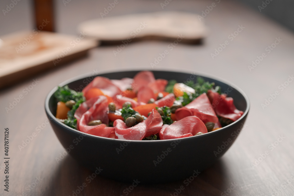 Salad with kale, bresaola and cherry tomatoes decorated with capers in black bowl on walnut wood table Salad with kale, bresaola and cherry tomatoes decorated with capers in black bowl on walnut wood table