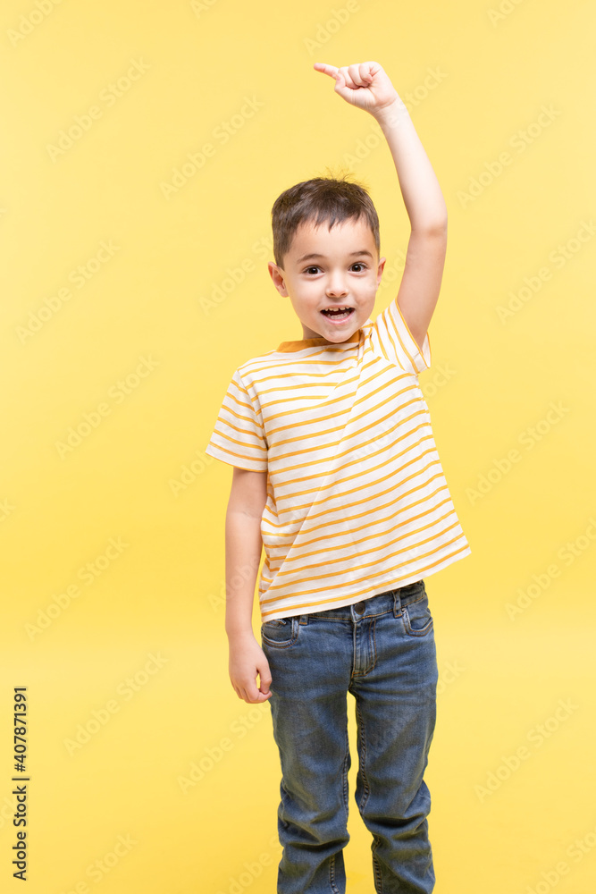 Child on a yellow studio background makes a gesture, shows the direction with his index finger, raising his hand above his head. T-shirt with white and yellow stripes, jeans. Emotional, positive face. Child on a yellow studio background makes a gesture, shows the direction with his index finger, raising his hand above his head. T-shirt with white and yellow stripes, jeans. Emotional, positive face.