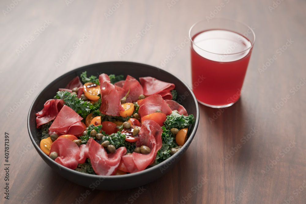 Salad with kale, bresaola and cherry tomatoes decorated with capers in black bowl and glass of red drink on walnut wood table Salad with kale, bresaola and cherry tomatoes decorated with capers in black bowl and glass of red drink on walnut wood table