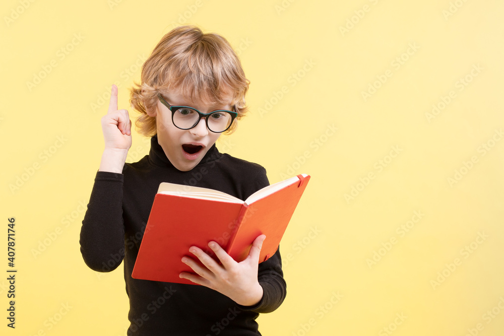 Blond Caucasian schoolboy surprised, inspired by an idea, wearing glasses. Raised index finger up, holding red notepad, wearing black sweater. Yellow background, copy space. Positive emotions on face. Blond Caucasian schoolboy surprised, inspired by an idea, wearing glasses. Raised index finger up, holding red notepad, wearing black sweater. Yellow background, copy space. Positive emotions on face.