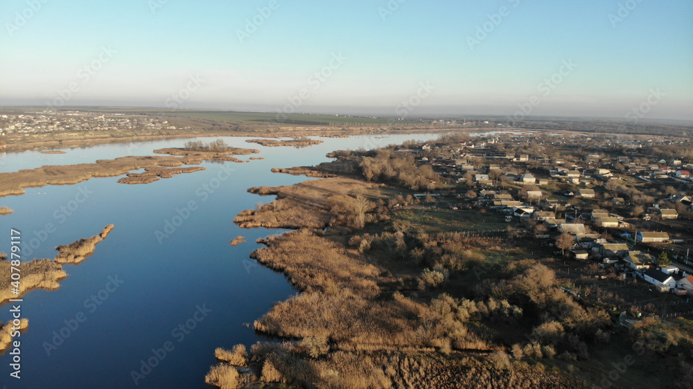 The estuary of a river with blue water. There are dry grass, reeds and trees on the bank and in the middle of the river. There are village with residential houses on the shore.
