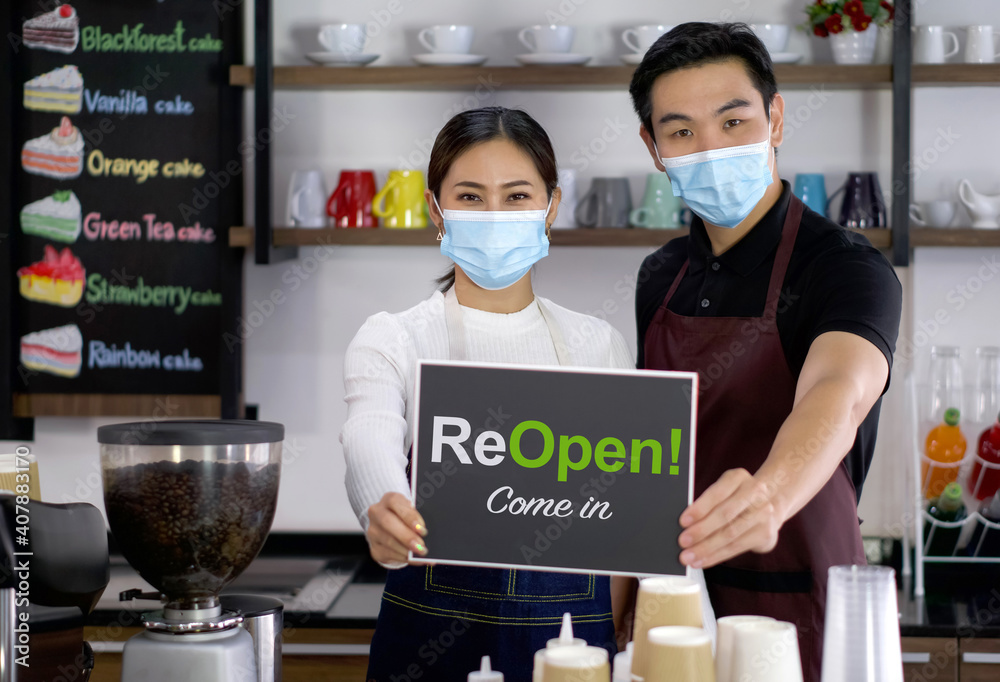 The shopkeeper and barista, wearing a face mask, holds an ReOpen! sign in front of a coffee shop counter. Morning atmosphere in a coffee shop. The shopkeeper and barista, wearing a face mask, holds an ReOpen! sign in front of a coffee shop counter. Morning atmosphere in a coffee shop.