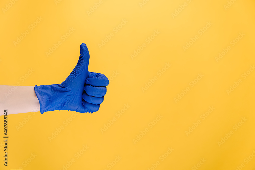 Close up of male in medical protective blue gloves makes thumb up gesture, demonstrates approval or agreement, isolated on yellow wall with copy space for advertisement. Hand sign. Like gesture