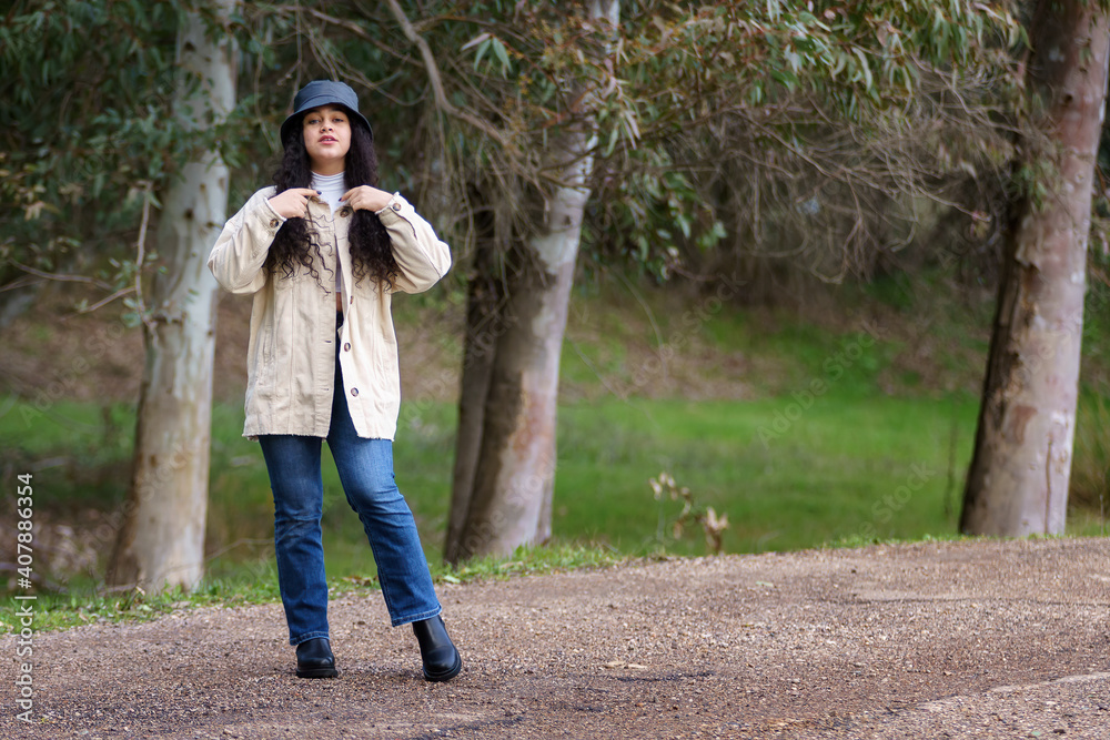 Fototapeta premium Chica adolescente hispana de pelo negro y rizado en el camino en el bosque en invierno con abrigo y sombrero.