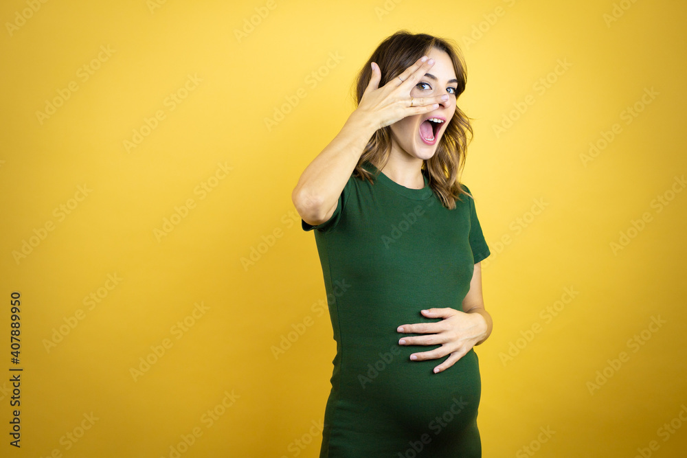 Young beautiful brunette woman pregnant expecting baby over isolated yellow background peeking in shock covering face and eyes with hand, looking through fingers with embarrassed expression Young beautiful brunette woman pregnant expecting baby over isolated yellow background peeking in shock covering face and eyes with hand, looking through fingers with embarrassed expression