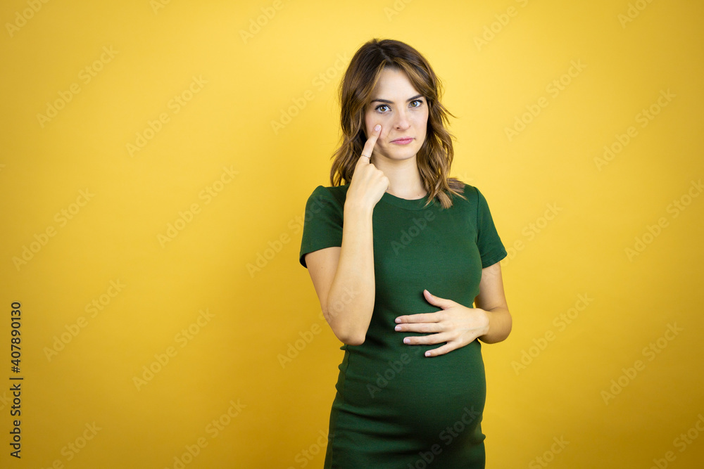 Young beautiful brunette woman pregnant expecting baby over isolated yellow background Pointing to the eye watching you gesture, suspicious expression Young beautiful brunette woman pregnant expecting baby over isolated yellow background Pointing to the eye watching you gesture, suspicious expression